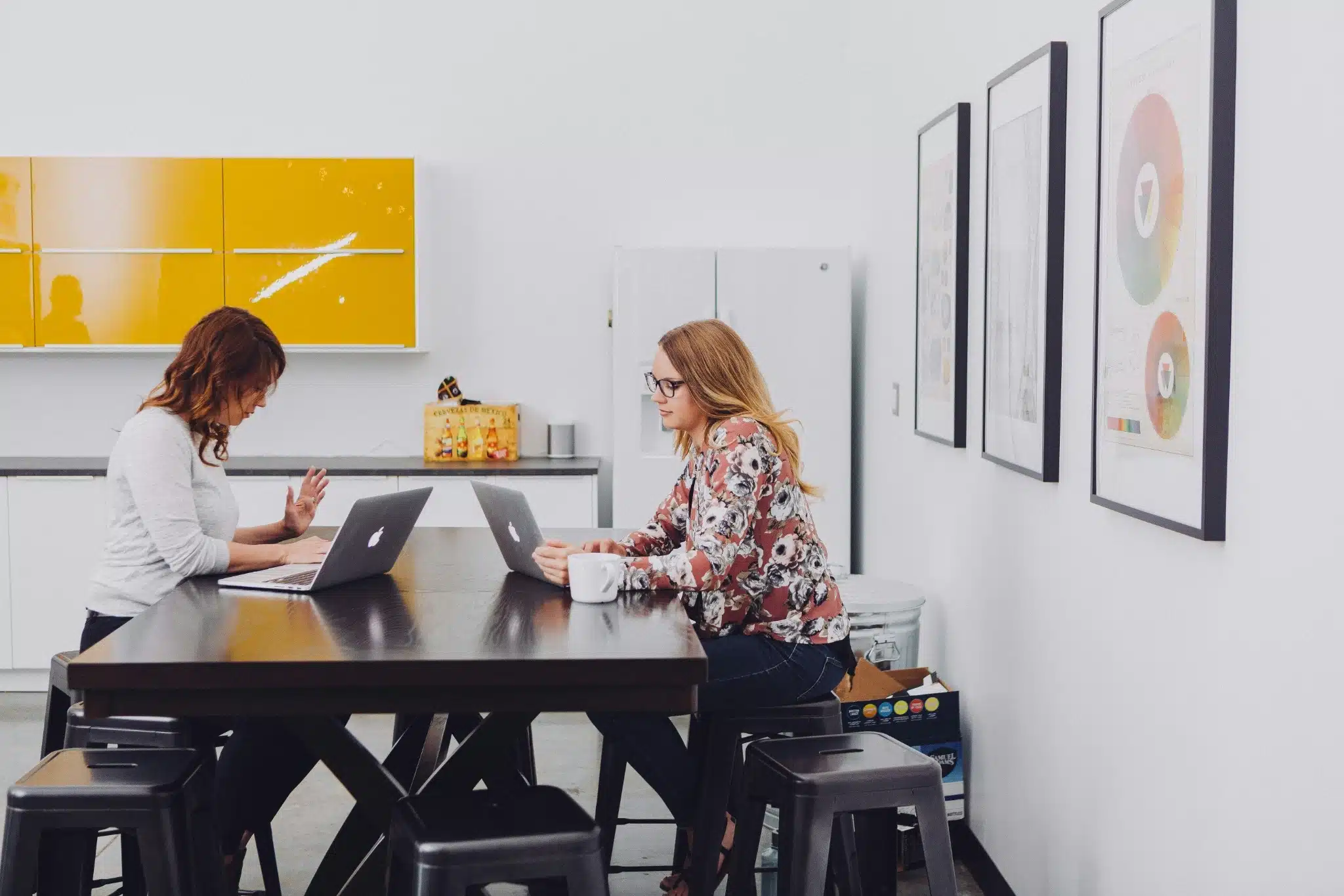 Two women sit at a black table working on laptops in a modern office kitchen with yellow cabinets and framed charts on the white walls. Focused on their work, they discuss community charity marketing strategies over coffee.