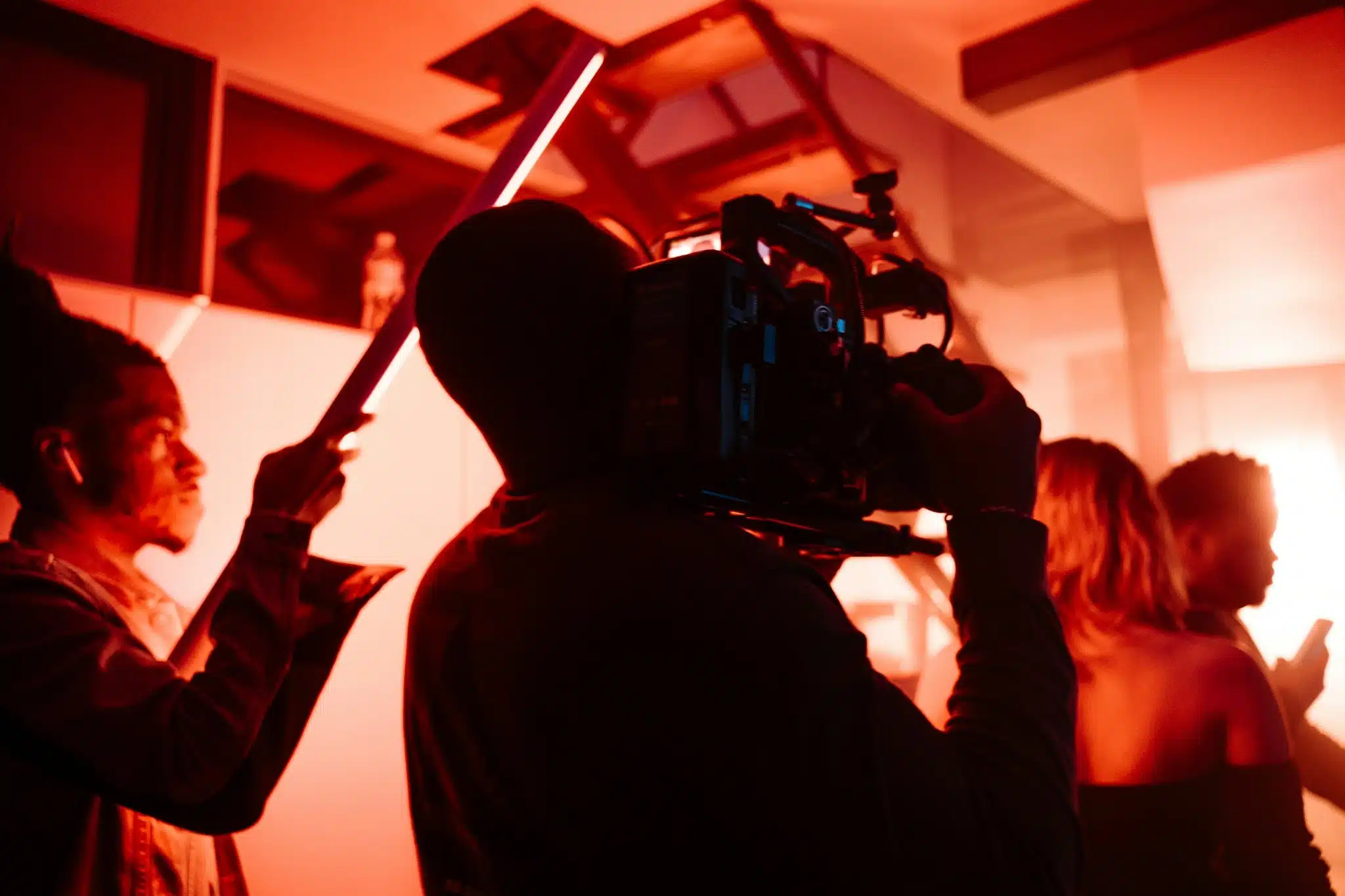 A camera operator films a scene with three people under dramatic red lighting, capturing a vivid moment that could power Community Charity Marketing. One person holds a light stick, while geometric shapes and intense illumination fill the background.