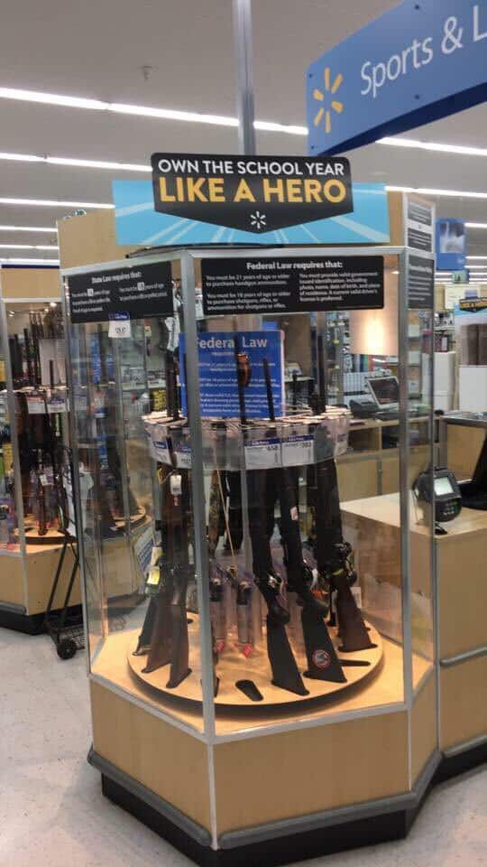 Employee mistakenly places display sign "Own the School like a Hero" above a gun display stand implying Walmart supports school shootings.
