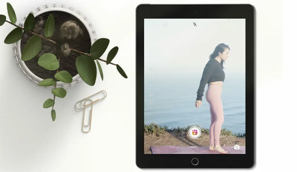 Flat lay of tablet showcasing an Instagram Reel of a woman by the sea. To the left-hand side, a plant and paperclips on a white background. Instagram search and explore