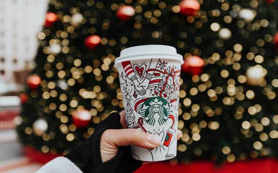 A woman holding a christmas-themed Starbucks cup in front of a lit up Christmas tree