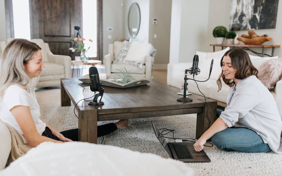Two women sitting on the floor of a living room speaking into microphones to record a podcast.