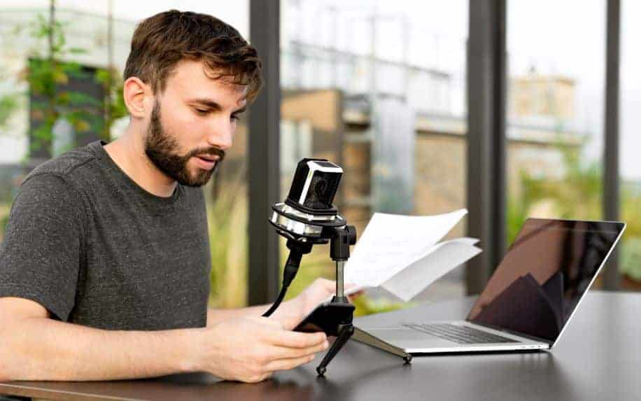 A man reading a script off of his phone into a microphone. The mic is connected to his Macbook.