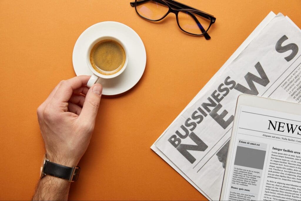 Business News Articles-Cropped view of man holding cup with coffee near glasses and business newspaper on orange