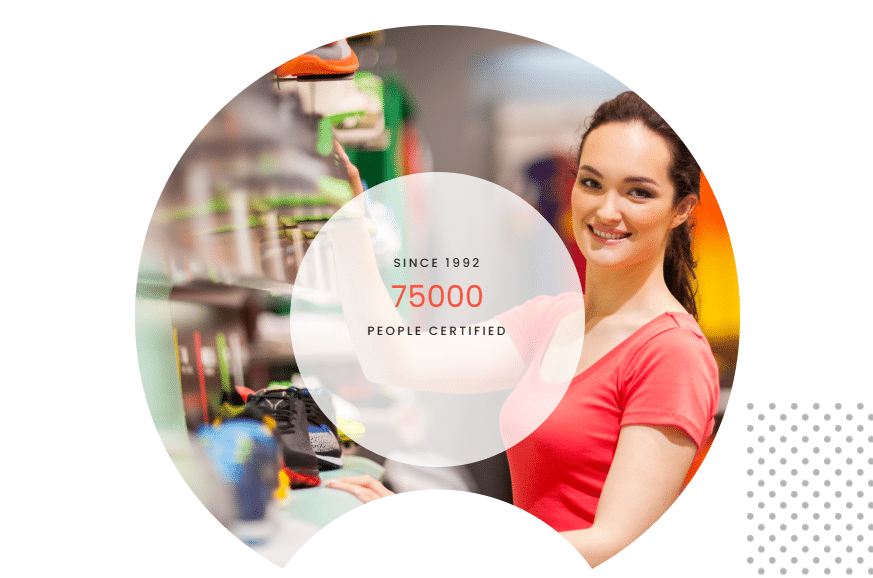 A smiling woman in a coral shirt arranges shoes on a shop shelf. Text overlay reads: “Since 1992, 75,000 people certified.” The scene highlights the impact of expert training provider support amid colourful merchandise. Training Website Design