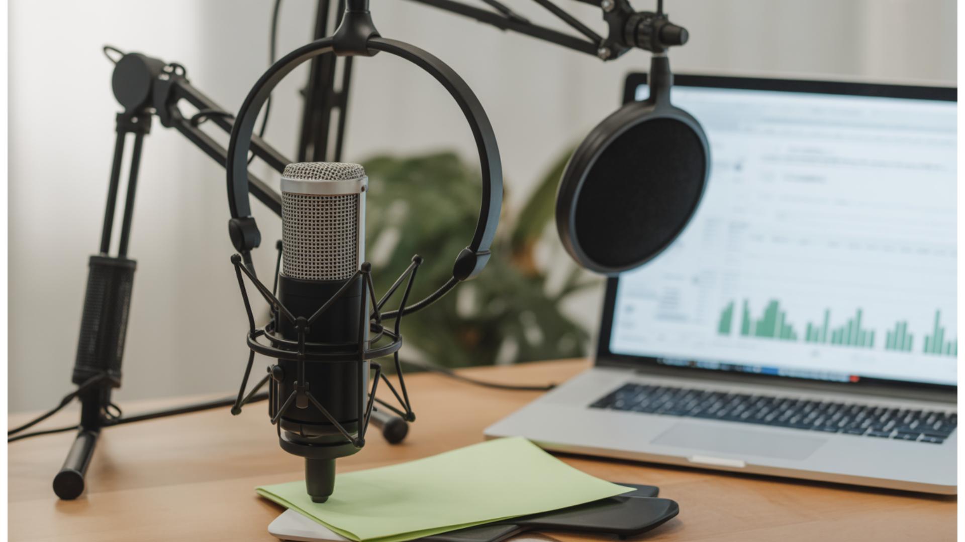 A podcast recording setup with a microphone on an adjustable arm, pop filter, headphones, and a laptop displaying charts—everything you need to create podcasts for your business—neatly arranged on a wooden desk with papers and a mobile phone.
