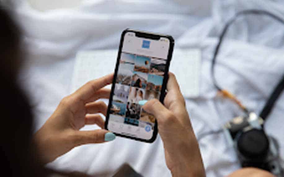 Influencer Marketing: A person with light blue nail polish uses a smartphone to browse a photo gallery, possibly curating content for influencer marketing; a camera and headphones are visible in the blurred background.