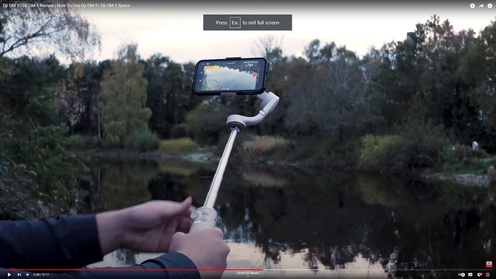 A person holds a smartphone mounted on a DJI Osmo 5 gimbal stabilizer, filming a calm river surrounded by trees and foliage at dusk. The phone screen displays the serene scene being recorded.
