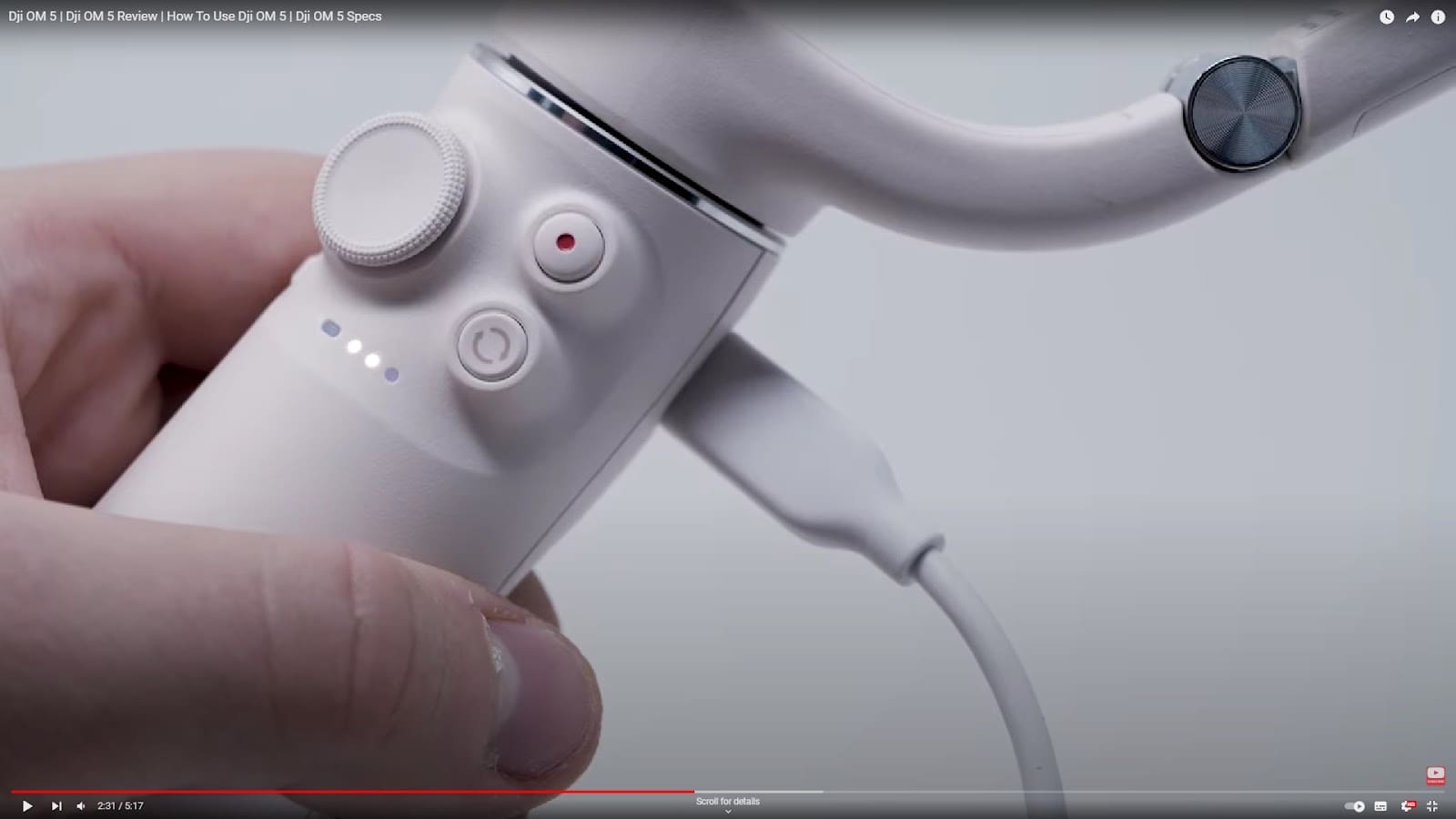 DJI Osmo 5 Review: A close-up of a hand holding a white DJI Osmo 5 handheld device with buttons, indicator lights, and a USB cable plugged in for charging. The device appears to be a gimbal or stabilizer.